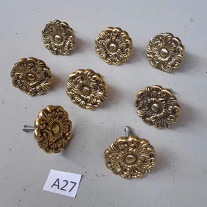May include: A collection of eight antique brass cabinet knobs with a floral design. The knobs are round with intricate detailing and a warm, golden hue. The image includes a small white tag with the text "A27".