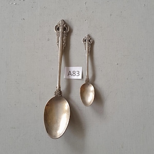 May include: Two antique silver spoons with decorative handles and oval bowls are shown against a muted grey backdrop. The larger spoon is approximately 20 cm long, while the smaller spoon is about 13 cm long. A small white tag with the text "A83" is placed between the spoons.