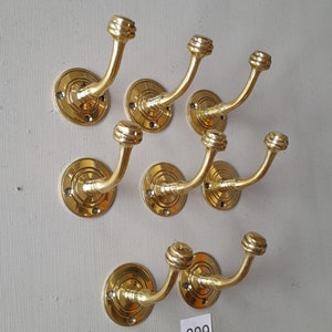 May include: A collection of seven brass coat hooks with a decorative, rounded top and circular base. The hooks are mounted on a light gray wall, with a small pile of brass screws below. The number "999" is on a small white label.
