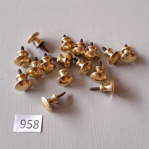 Könnte beinhalten: Eine Ansammlung kleiner, glänzender Messingknöpfe mit Schrauben auf einer weißen Oberfläche. Die Knöpfe haben eine abgerundete, pilzartige Form. Ein kleines weißes Etikett zeigt die Nummer "958".