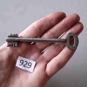 May include: A silver key with a round head and a long, thin shaft. The key has a small, intricate design on the top and the word "UPS" engraved on the head. The key is being held in a hand, and a white piece of paper with the number "929" written on it is visible in the background.