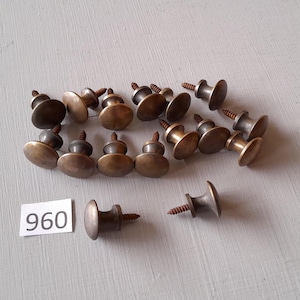 May include: A group of 16 antique brass cabinet knobs with screws. The knobs are round with a raised center and a slightly domed top. The screws are silver in color. The number 960 is written on a white piece of paper.