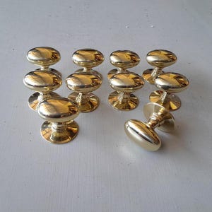 Puede incluir: Un conjunto de diez pomos de puerta de latón pulido. Cada pomo tiene forma ovalada y base redonda. Los pomos están dispuestos sobre una superficie gris claro, reflejando la luz ambiental.