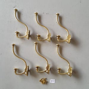 May include: Six vintage brass coat hooks with a curved design, arranged on a light gray surface. Each hook features a decorative base and a curved arm ending in a rounded top. A small decorative piece and a tag with "A92" are also present.