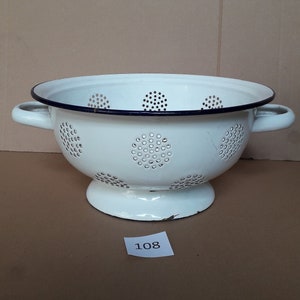 May include: White enamel colander with a blue rim and a pattern of small circles. The colander has two handles and a pedestal base.