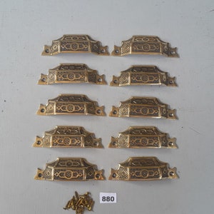 May include: Ten gold-toned cabinet pulls with an ornate design. The pulls are arranged in two rows of five. The pulls are accompanied by a small bag of screws.