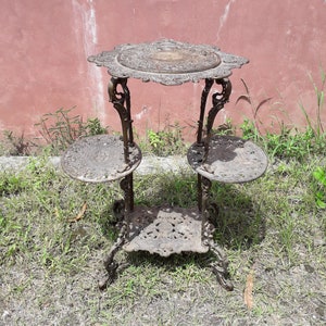 Puede incluir: Un soporte para plantas de hierro fundido de tres niveles con intrincados diseños florales. El soporte tiene una parte superior redonda y tres patas. El soporte está pintado de negro y tiene un acabado desgastado.