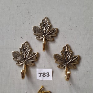 Puede incluir: Tres ganchos de hoja de color dorado con un gancho en la parte inferior de cada hoja. Los ganchos están sobre una superficie blanca con un pequeño trozo de papel en el que está escrito el número 783. También hay un montón de tornillos dorados en la superficie.