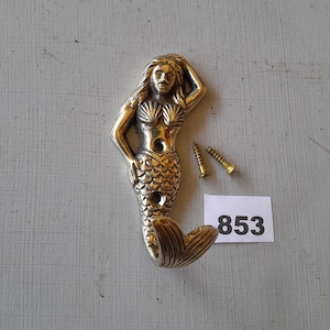 May include: Silver mermaid wall hook with a tail that forms a hook. The mermaid is wearing a seashell bra and has a detailed face. Two screws are included. The number 853 is written on a white piece of paper.