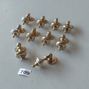 May include: Twelve gold-toned cabinet knobs with a decorative design. The knobs have a round base and a raised, ornate top. One knob is angled to the side.