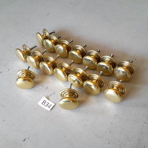 May include: A collection of fourteen round, polished brass cabinet knobs, each featuring a smooth, domed head and a short metal mounting screw extending from the back. A small white rectangular tag with the black printed text B34 is included.