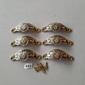 May include: Six ornate brass drawer pulls with a leaf design. The pulls are arranged in two rows of three. The pulls are accompanied by a small bag of screws labeled "823".