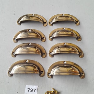 May include: Eight brass-colored, half-moon drawer pulls with screw holes. The pulls are arranged in two rows of four. A small white label with the number "797" is visible, along with a pile of brass screws.