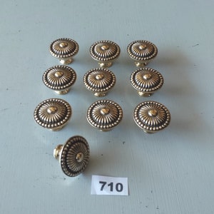 Puede incluir: Doce pomos de armario de color dorado con un diseño circular decorativo en relieve. Los pomos tienen un borde con cuentas y un centro elevado. Un pomo se muestra por separado. El número 710 está escrito en una etiqueta blanca.
