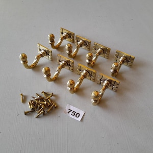 Peut inclure: Huit crochets dorés avec un design en spirale décoratif et un sommet arrondi. Les crochets sont fixés à des plaques arrière carrées. Un tas de petites vis dorées est inclus. Le nombre 750 est écrit sur une étiquette blanche.