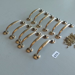 May include: A set of twelve brass-toned cabinet handles with a curved design, arranged in rows on a gray surface. Small brass screws are scattered nearby. The handles have a classic, elegant appearance, suitable for furniture restoration or new builds.