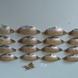 May include: A set of 15 gold-toned metal cabinet pulls with a scalloped design. The pulls are arranged in three rows of five. A small pile of screws is included.