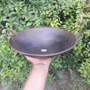 May include: A large, black cast iron wok with two handles. The wok is slightly curved and has a rough, textured surface.