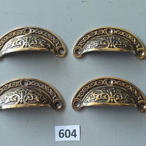May include: Four antique brass drawer pulls with ornate floral designs. Each pull is shaped like a half-circle and has two small holes for mounting.