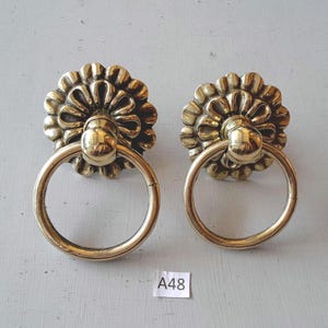May include: Two brass door pulls with a floral design and ring handles. The pulls have a warm, golden hue and are mounted on a light gray surface. The tag in the image reads "A48".