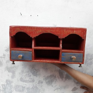 May include: A rustic, red wooden wall shelf with three arched compartments and two blue drawers with brass knobs. The shelf has a distressed finish, giving it a vintage look. The shelf is supported by small metal feet.