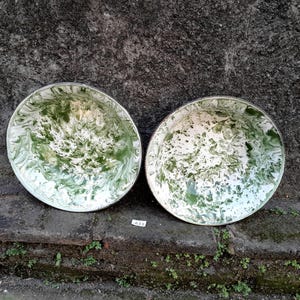 May include: Two round enamelware bowls with a white and green marbled design. The bowls have a silver rim and are set against a textured, dark grey background. The bowls are sitting on a brick surface. The text "A33" is visible.