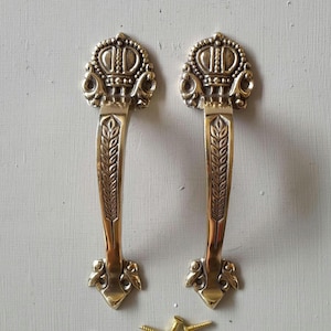May include: Two ornate brass door handles. Each handle features a crown design at the top and a wheat stalk pattern down the center. The handles are mounted on a light gray surface, with a few brass screws visible at the bottom.