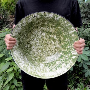 May include: Large, round enamelware bowl with a green and white marbled pattern. The bowl has a black rim and is held by a person. The bowl's interior features a swirling design, creating a unique and decorative piece.