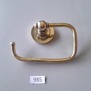 May include: A gold-toned toilet paper holder with a round base and a curved arm. The holder has a number "985" written on a white tag below it.