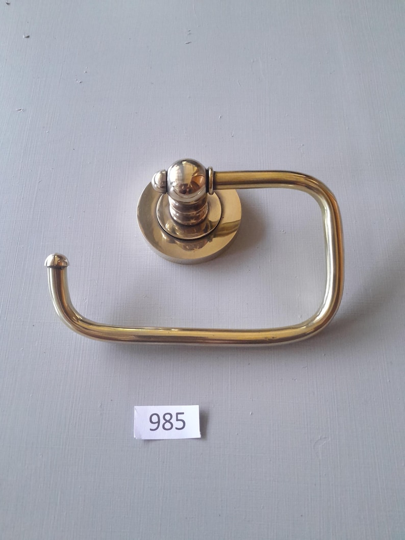 May include: A gold-toned toilet paper holder with a round base and a curved arm. The holder has a number "985" written on a white tag below it.