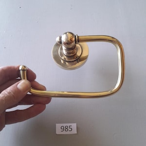 May include: A gold-toned metal toilet paper holder with a square base and a rounded top. The holder is designed to be mounted on a wall.