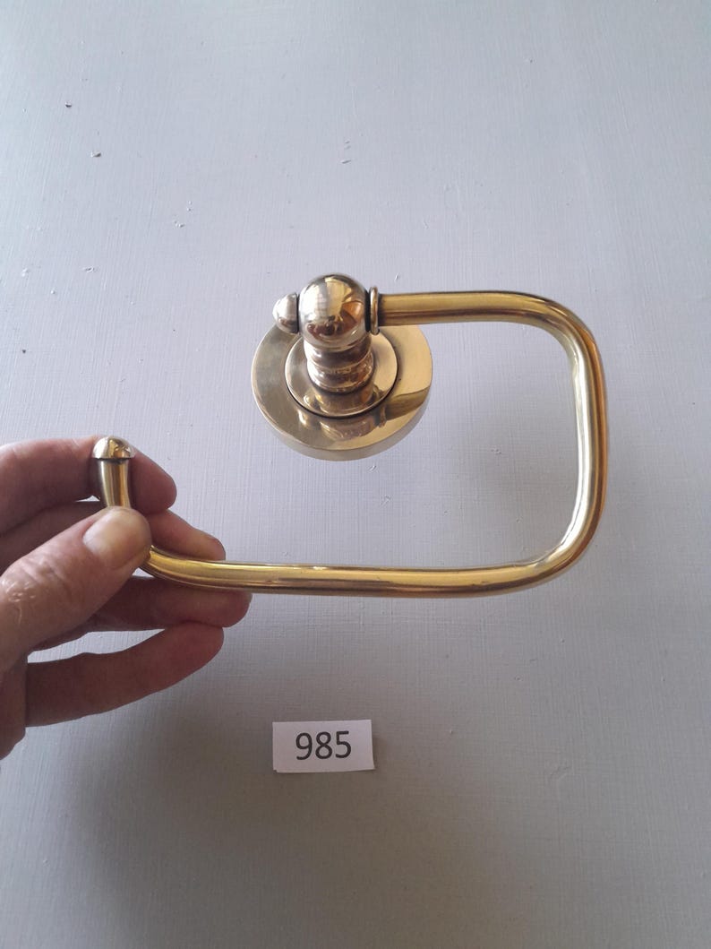 May include: A gold-toned metal toilet paper holder with a square base and a rounded top. The holder is designed to be mounted on a wall.
