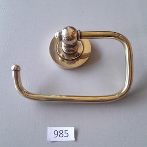 May include: A gold-toned toilet paper holder with a round base and a curved arm. The holder has a decorative knob on the top. The number 985 is written on a white label.