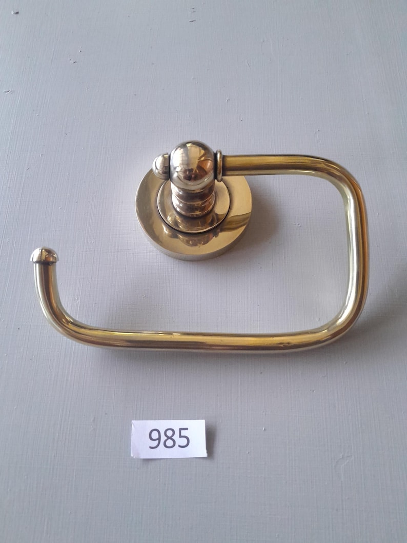 May include: A gold-toned toilet paper holder with a round base and a curved arm. The holder has a decorative knob on the top. The number 985 is written on a white label.