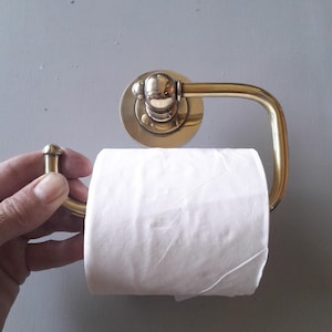 May include: A brass toilet paper holder with a round base and a curved arm. A roll of white toilet paper is on the holder.