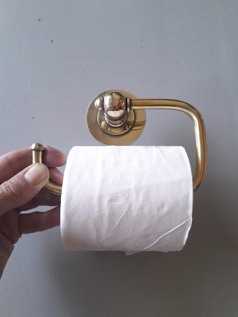 May include: A brass toilet paper holder with a round base and a curved arm. A roll of white toilet paper is on the holder.