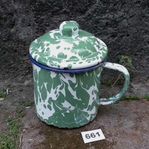 Pode incluir: Uma caneca de esmalte branco com um padrão de respingos verdes e uma tampa correspondente. A caneca tem uma borda e uma alça azuis. O número "661" está em uma etiqueta branca na imagem.