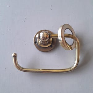 May include: A brass toilet paper holder with a round base and a ring-shaped arm. The holder is mounted on a wall.