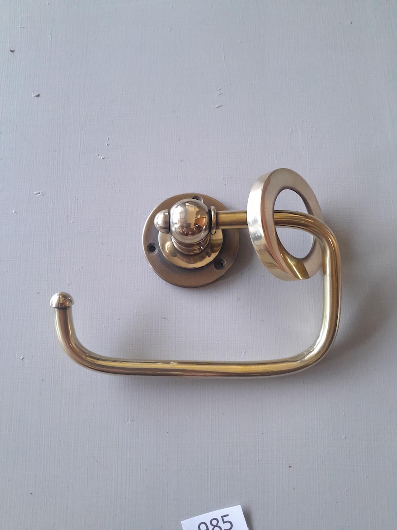 May include: A brass toilet paper holder with a round base and a ring-shaped arm. The holder is mounted on a wall.