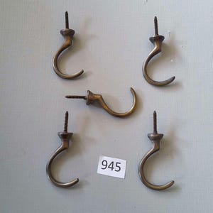 Peut inclure: Cinq crochets en laiton antique avec un dos à vis. Les crochets sont tous du même design, avec une forme incurvée et un petit bouton en haut. Les crochets sont disposés en demi-cercle sur une surface bleu clair. Le numéro 945 est imprimé sur une étiquette blanche.