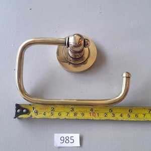 May include: A brass toilet paper holder with a round base and a curved arm. The holder is mounted on a white wall. The measurement on the tape measure is 10 inches.