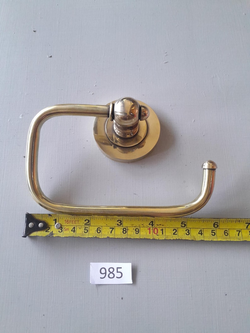 May include: A brass toilet paper holder with a round base and a curved arm. The holder is mounted on a white wall. The measurement on the tape measure is 10 inches.
