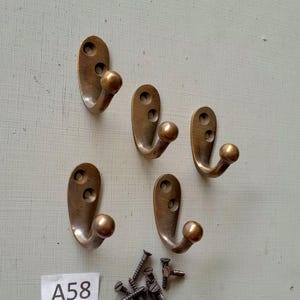 Op de afbeelding: Vijf antieke messing kapstokken met een afgeronde kogelvormige punt en twee schroefgaten. De haken zijn op een lichtgroene muur gemonteerd. De afbeelding bevat ook een kleine stapel schroeven en de tekst "A58".