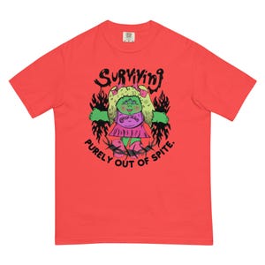 May include: A coral-colored short-sleeve t-shirt featuring a graphic of a green three-eyed doll with blonde hair wearing a pink dress. The text reads "SURVIVING" above the doll and "PURELY OUT OF SPITE." below, framed by black flames and barbed wire.