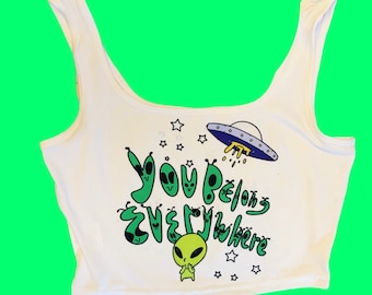 May include: White crop top with a green alien graphic and the text "You Belong Everywhere". The design includes a UFO, stars, and several cartoon aliens. The top has a scoop neck and is sleeveless.