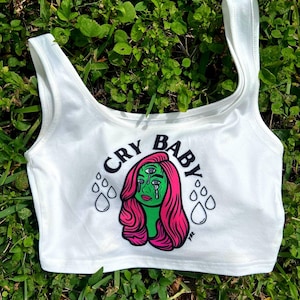 May include: White crop top with the text "CRY BABY" in a circular font. The graphic features a green-skinned woman with pink hair and three eyes, crying teardrops. The top is set against a background of green grass.
