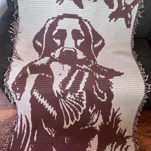 Puede incluir: Una manta rectangular de ganchillo con un diseño bicolor en marrón oscuro y beige. El patrón muestra un perro grande en el centro con tres patos volando encima. Los bordes de la manta están rematados con flecos de hilos sueltos.