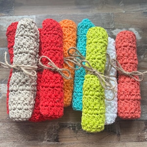 May include: A selection of rolled crochet washcloths in vibrant colours, tied with twine. The washcloths are in shades of red, orange, teal, and green. The crochet texture is visible, and the twine adds a rustic touch to the presentation.