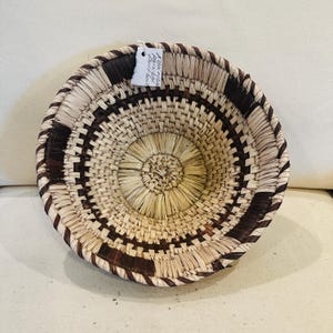 May include: A round woven basket with a natural beige and dark brown geometric pattern. The basket has a circular shape with a woven design. The rim and the pattern are dark brown. The center is a lighter beige color. A tag is attached to the basket.