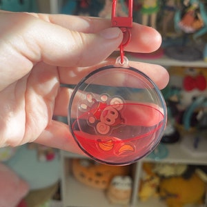 May include: A red and clear plastic keychain with a monkey and bananas inside. The monkey is wearing a red hat and is floating in red liquid.